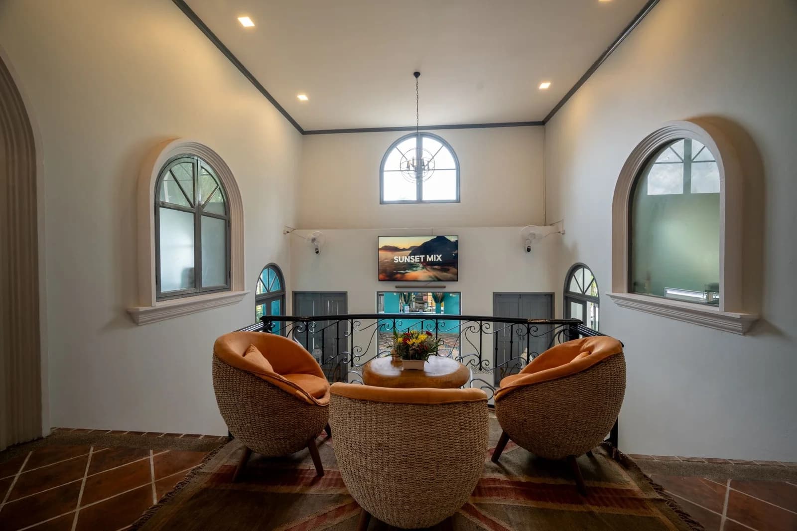 Upstairs lounge area inside Soho Clubhouse Phuket featuring rattan armchairs, warm lighting, and a balcony view overlooking the pool area and “Sunset Mix” display screen.
